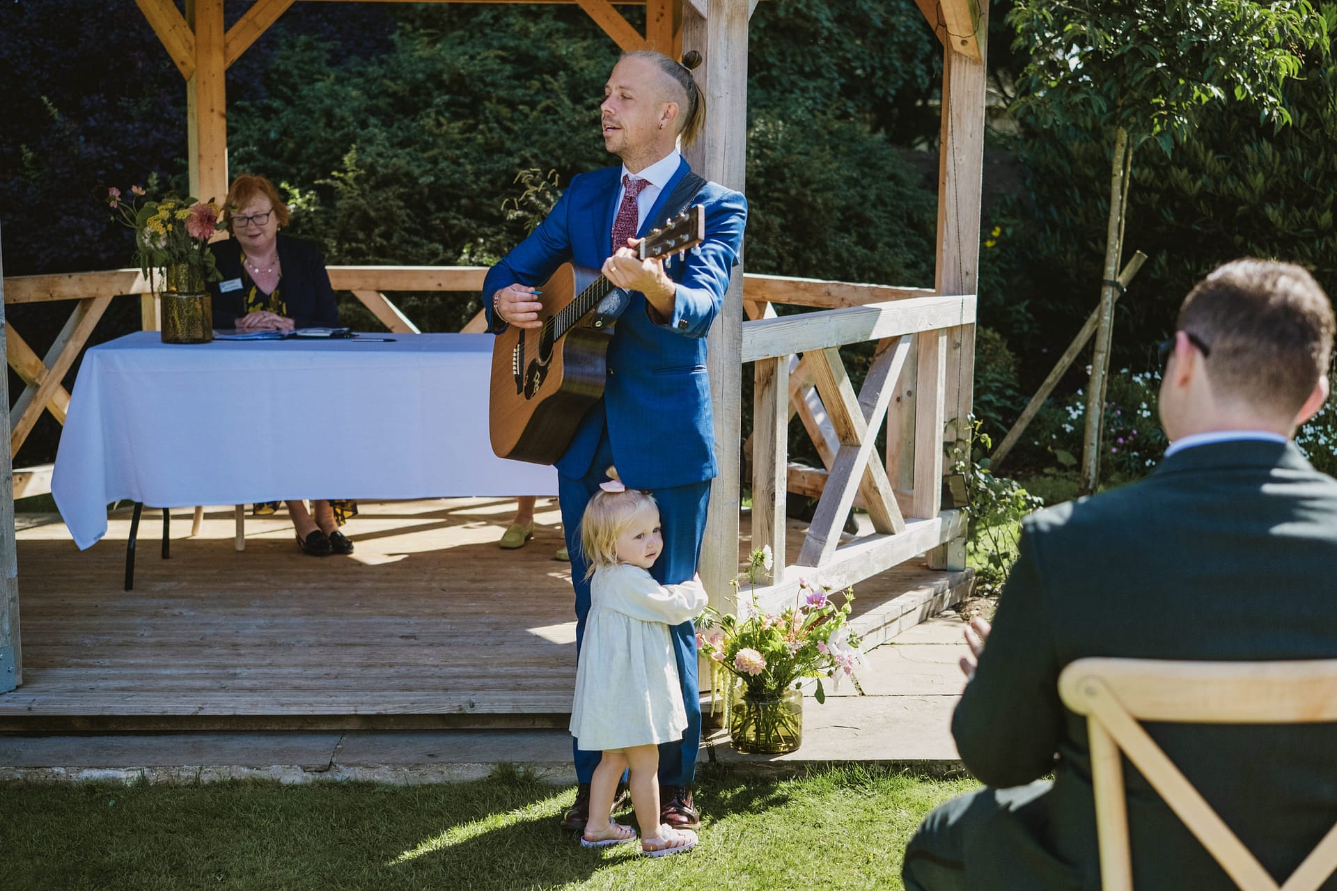 Man playing guitar outdoors, child hugging leg.