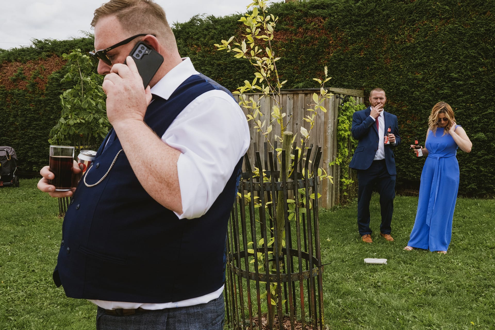 Man talks on phone, people with drinks in garden.