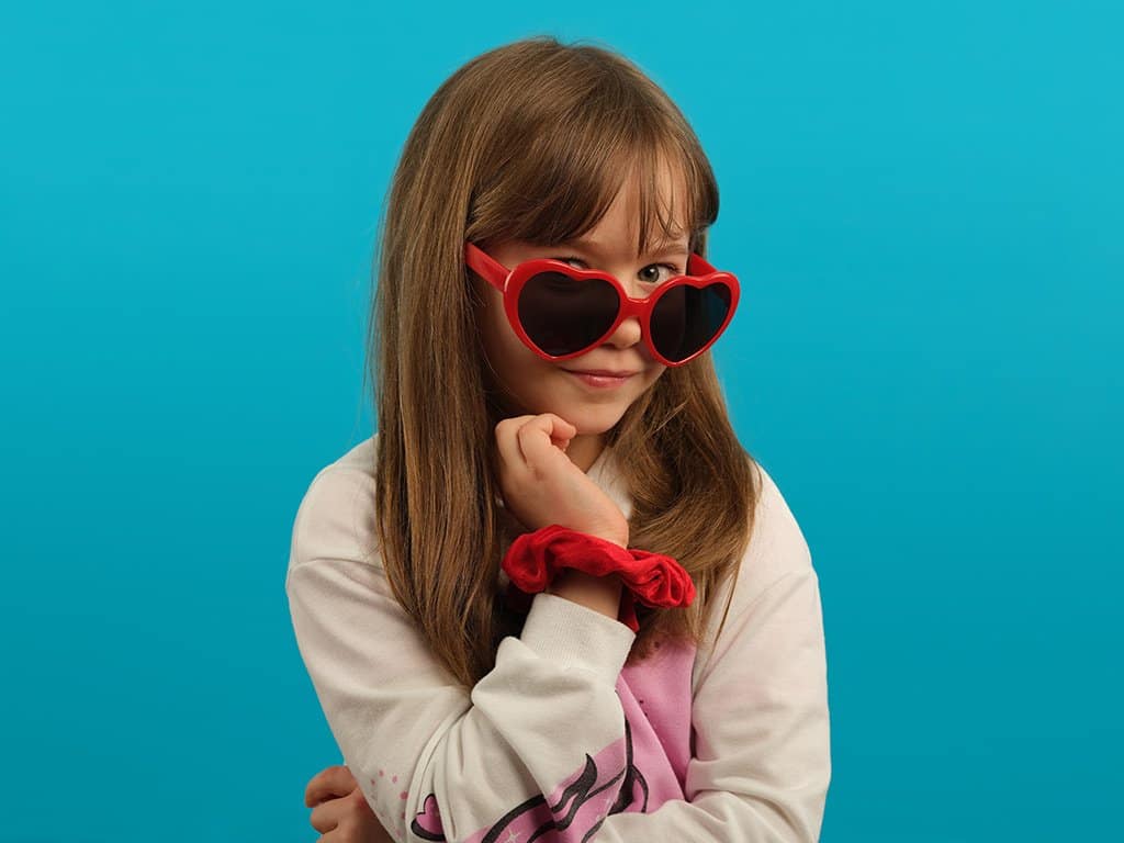 Studio portrait photography example - a young girl in heart-shaped sunglasses poses in front of a colourful blue background.