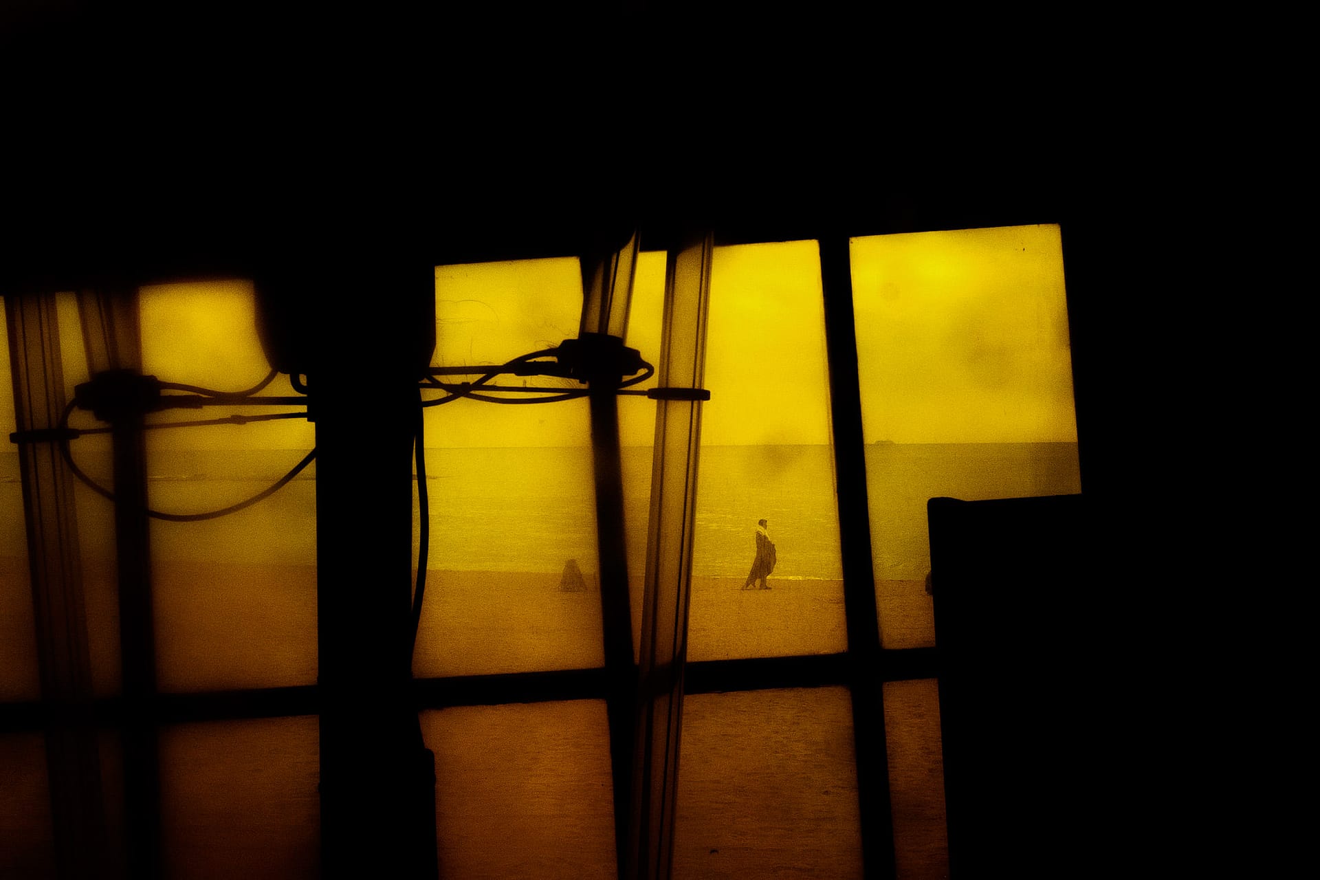 Street photograph captured in Barcelona, Spain. Figure is photographed in the distance on a beach, shot through a yellow window