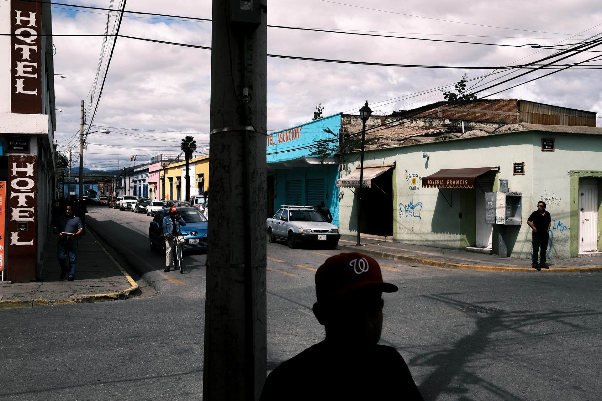Street photograph captured in Oaxaca, Mexico. Man in a baseball cap is silhouetted in the foreground, whilst we can see the characteristics of a Mexican street behind.