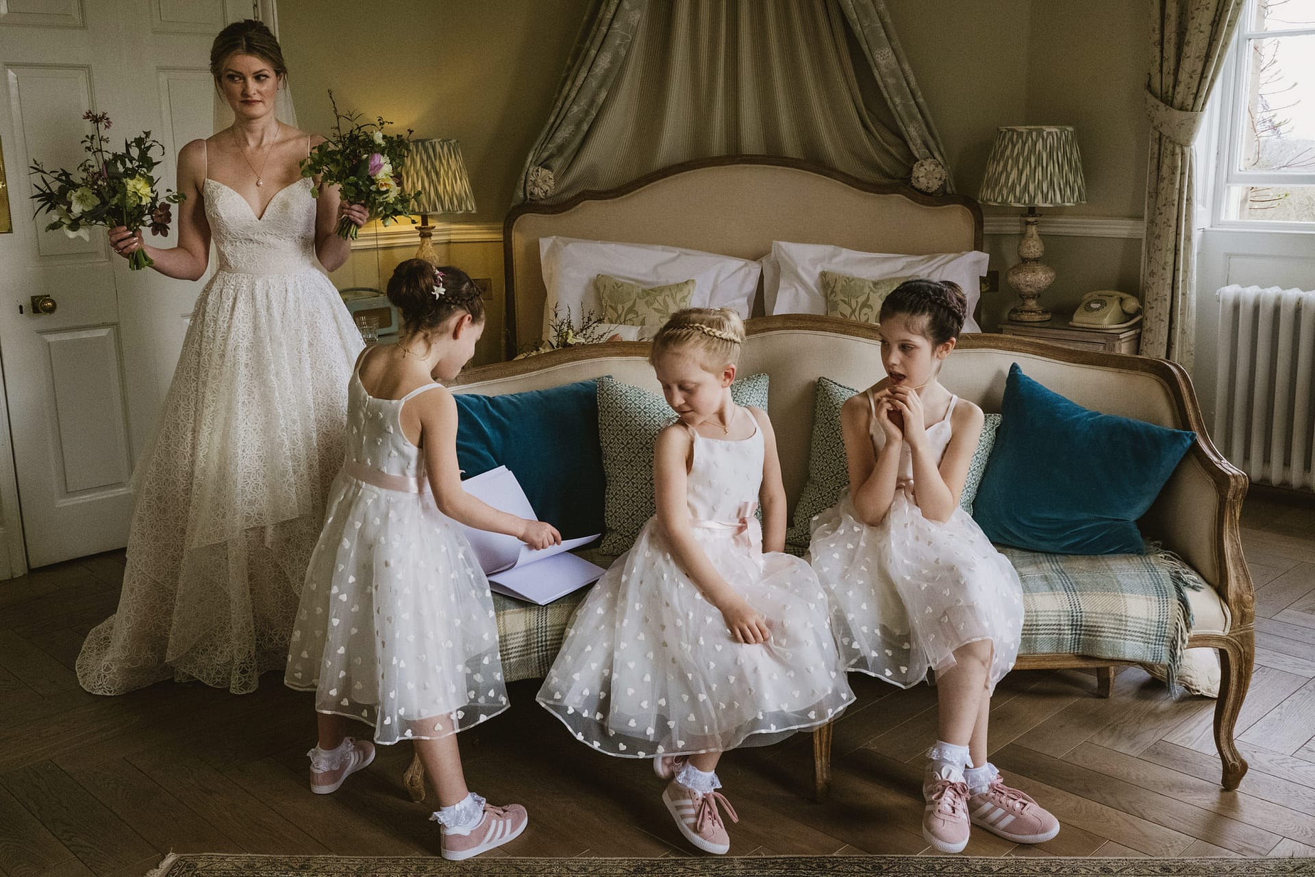 A wide, candid shot in a bright Middleton Lodge bridal suite. A bride in a lace gown stands holding two bouquets, acting as an anchor for three young flower girls in pink trainers who are seated on a vintage sofa, focused on a book during a quiet moment of pre-ceremony anticipation.