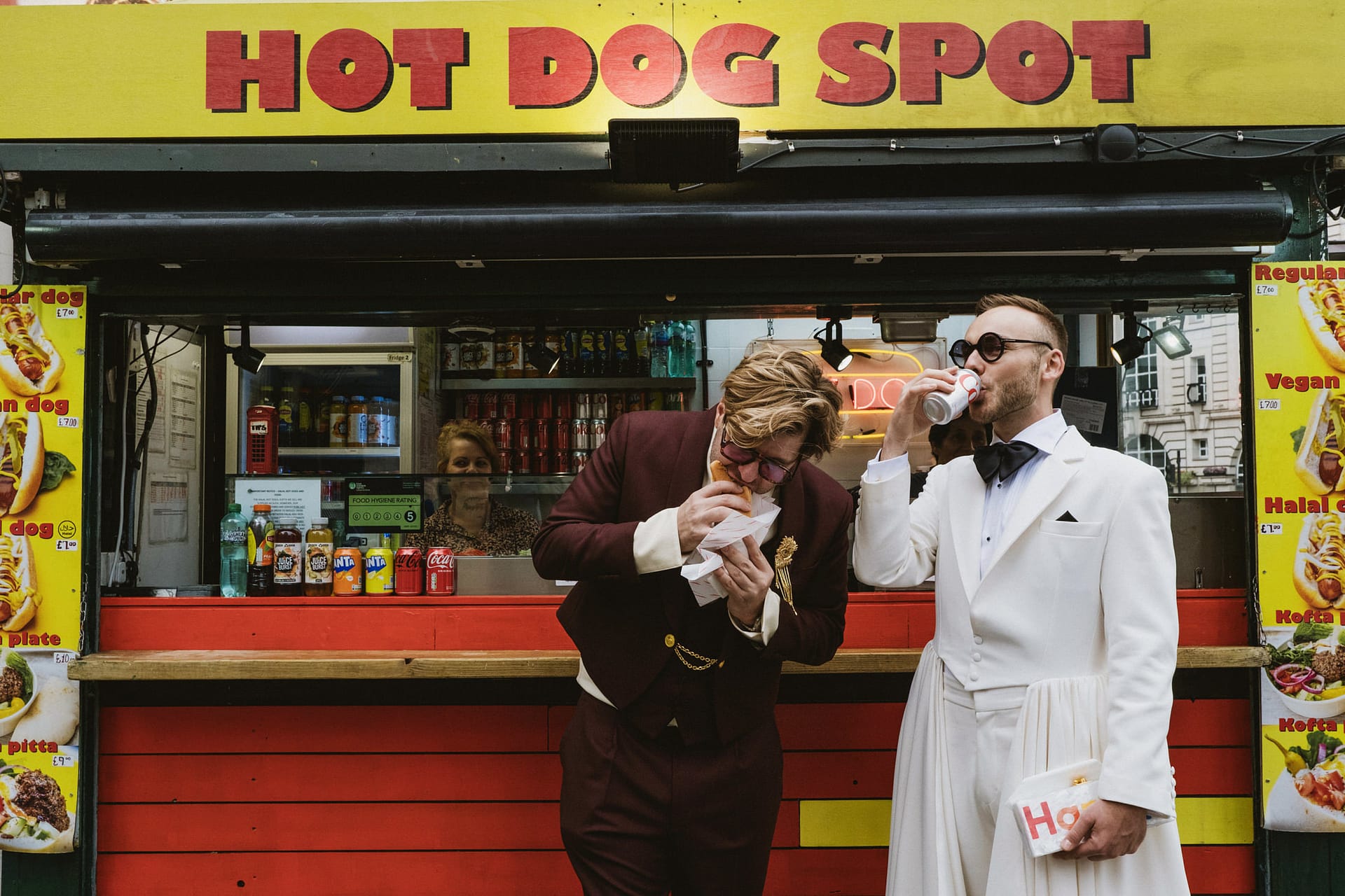 Two men in suits eating at hot dog stand.