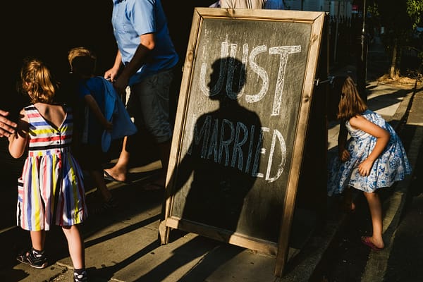 children are photographed either side of a chalkboard sign that reads "just married". The shadowed silhouette of another person sits over the text.