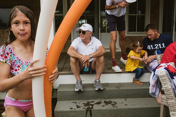 Documentary style family photography example: a girl stands facing the camera holding swimming pool floats to the left of the frame. To the right other members of the family sit and stand on the steps interacting with one another.