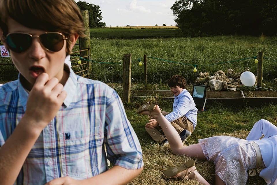 documentary wedding photography - kids play on haybales at an outdoor wedding reception. To the left a boy in aviator sunglasses looks towards the camera with a natural expression. To his left a boy sits on a haybale and on the far right of the frame we can see the legs of a girl clambering horizontally over another haybale