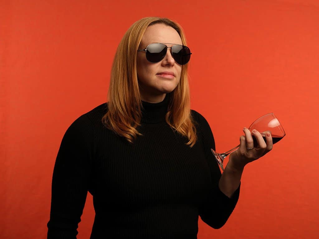 woman wearing sunglasses holding a wine glass filled with red wine on an orange background