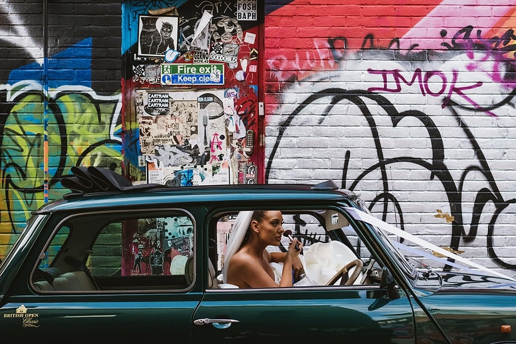 documentary style wedding photograph - bride touches up makeup in mini wedding car in front of a graffiti wall background on London street