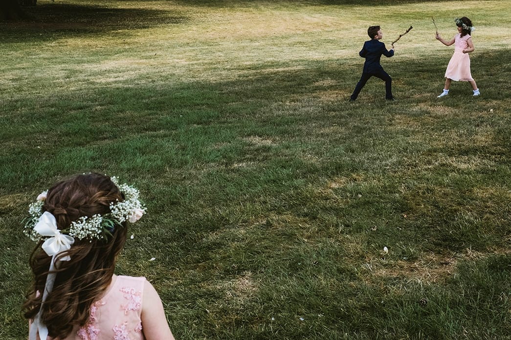 children at wedding reception have a sword fight whilst another flower girl watches on. Photographed in a reportage style