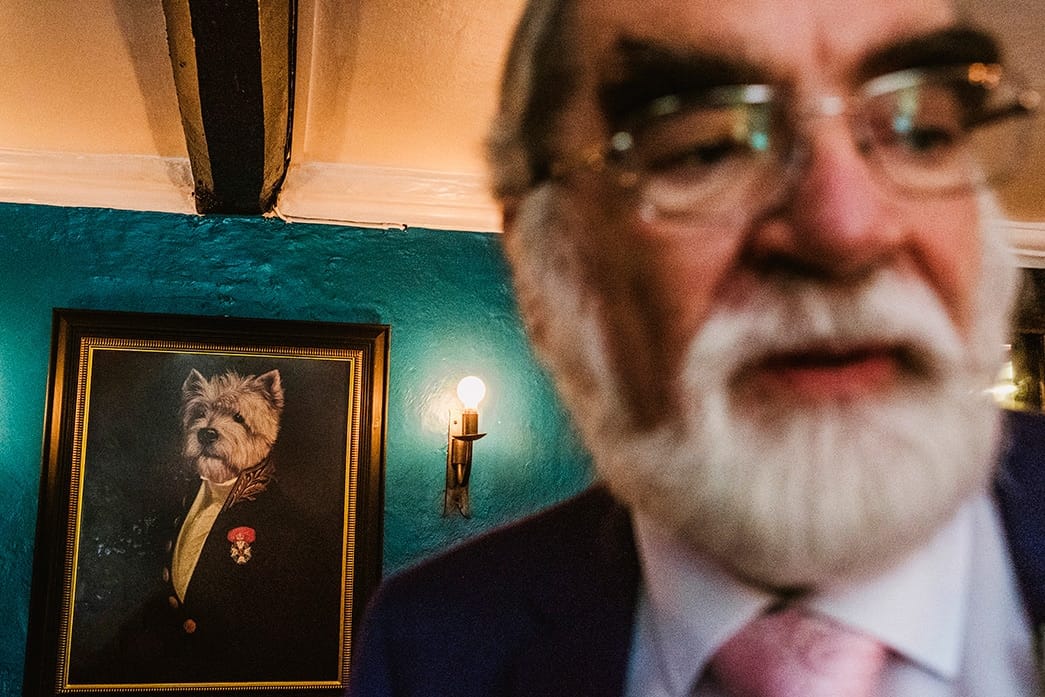 street style wedding image - man at wedding stands on right of frame, to his left is a portrait of a dog in a suit who humorously shares similar facial features.