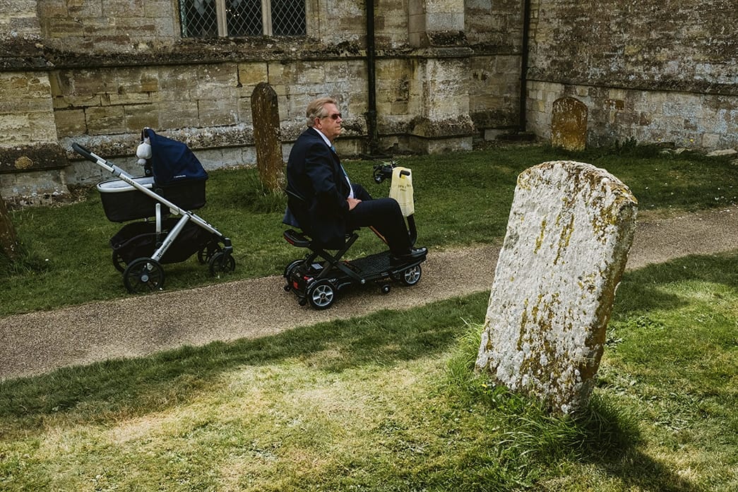 street style wedding image representing the journey of life - pram on left of frame, man on disability scooter in centre and gravestone to the right of the frame.