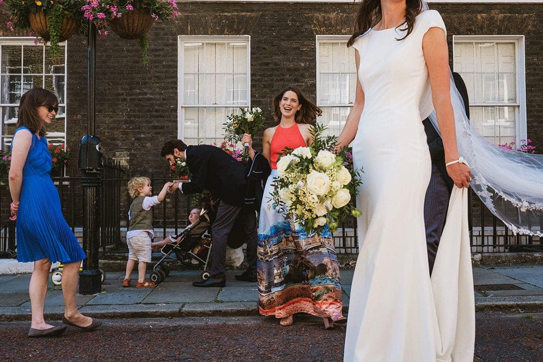 Documentary wedding photograph from an intimate wedding in London. In the foreground to the right of the frame a bride in white dress holds her wedding train, her veil floating out behind her, her face not included in the frame. In her right hand is her bridal bouquet, behind which a smiling bridesmaid holds another bouquet. Behind and to her right the groom bends down to take the hand of a young boy, a pram with another child framed between them. Far left of the frame a woman in a blue dress looks on.