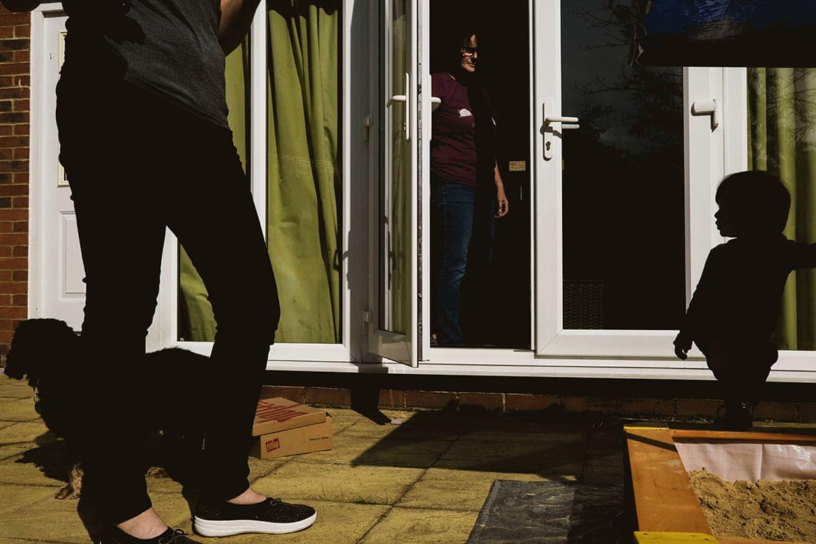 Family Photography outside a Yorkshire home. Centre (rear) a woman is framed in the doorway. Right of the frame a young boy is silhouetted behind a sandpit. Left of the frame we can see a woman's legs and a small silhouetted dog