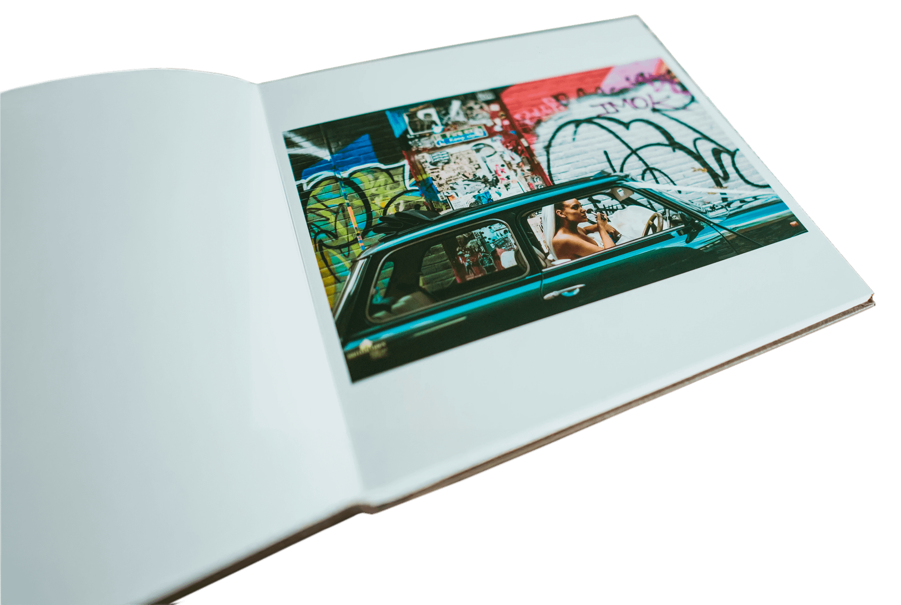 Wedding album of documentary style photographs. Page is open at a photograph of a bride applying her makeup in the front seat of a classic green Mini Cooper decked in wedding ribbons. Behind the car is a wall full of graffiti