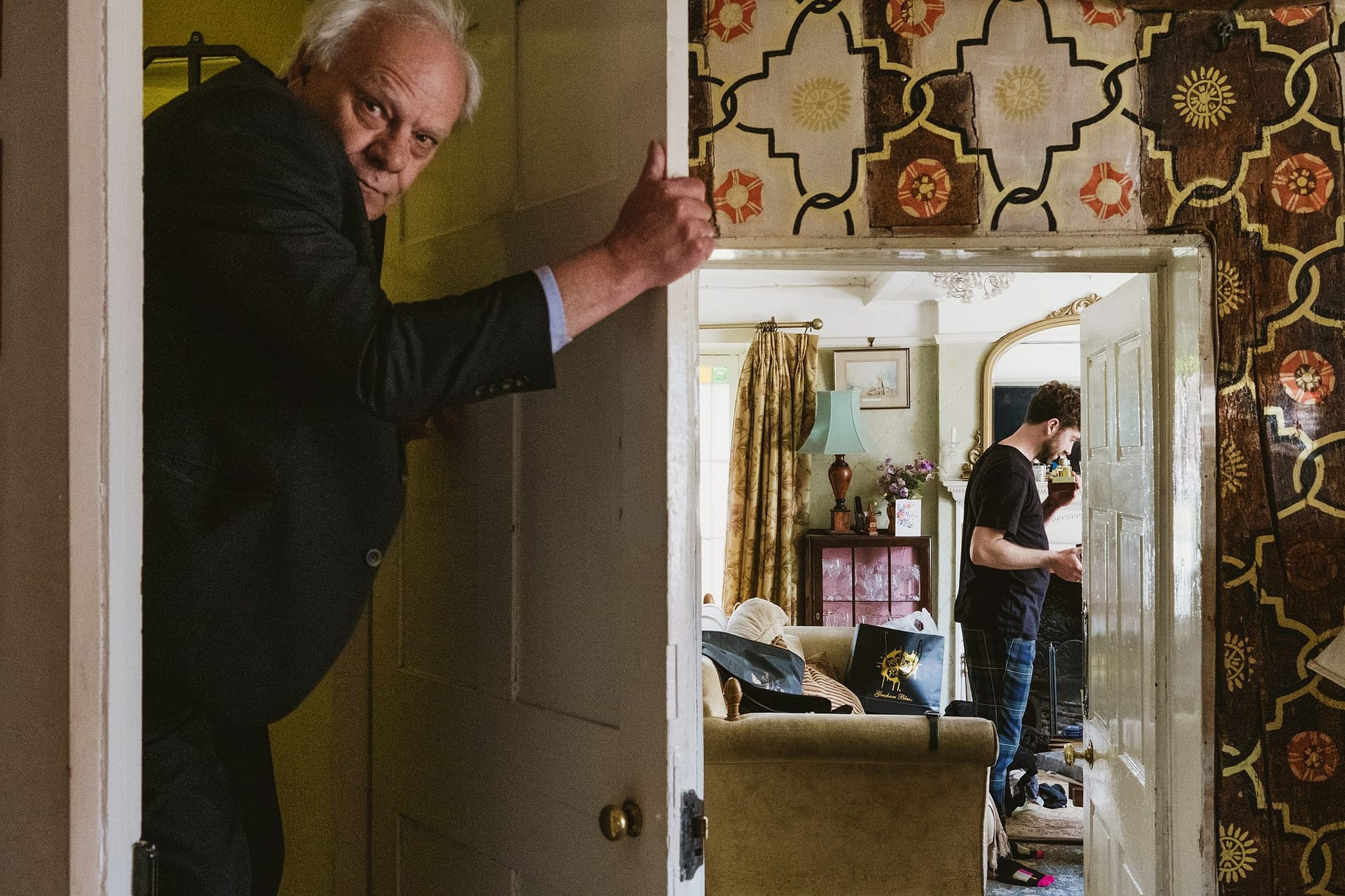 A man notices the camera as he walks into the room through a door on the left of the frame. On the right we can see through into another room where a groom prepares for his wedding