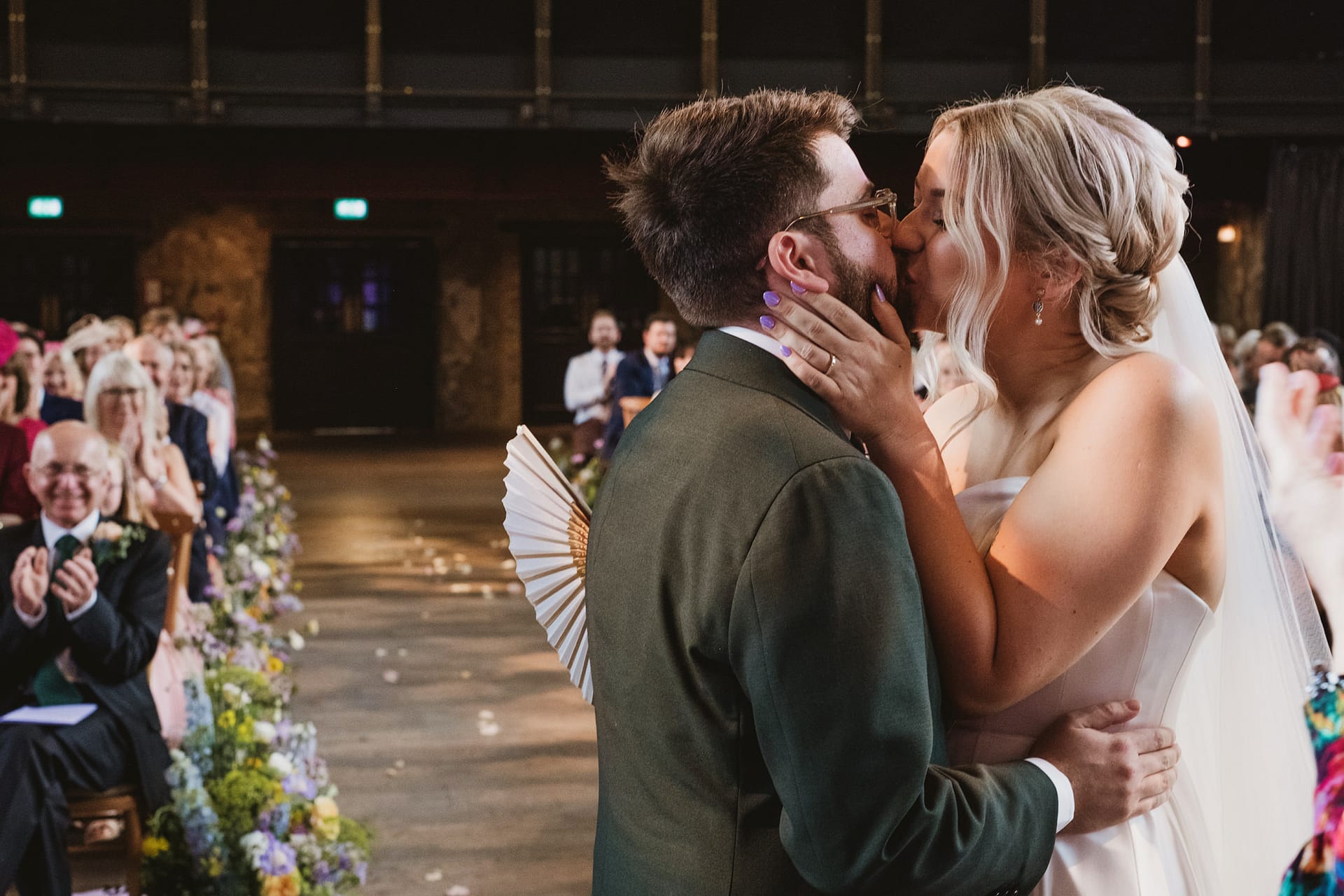 Bride and groom share wedding kiss, guests applauding.