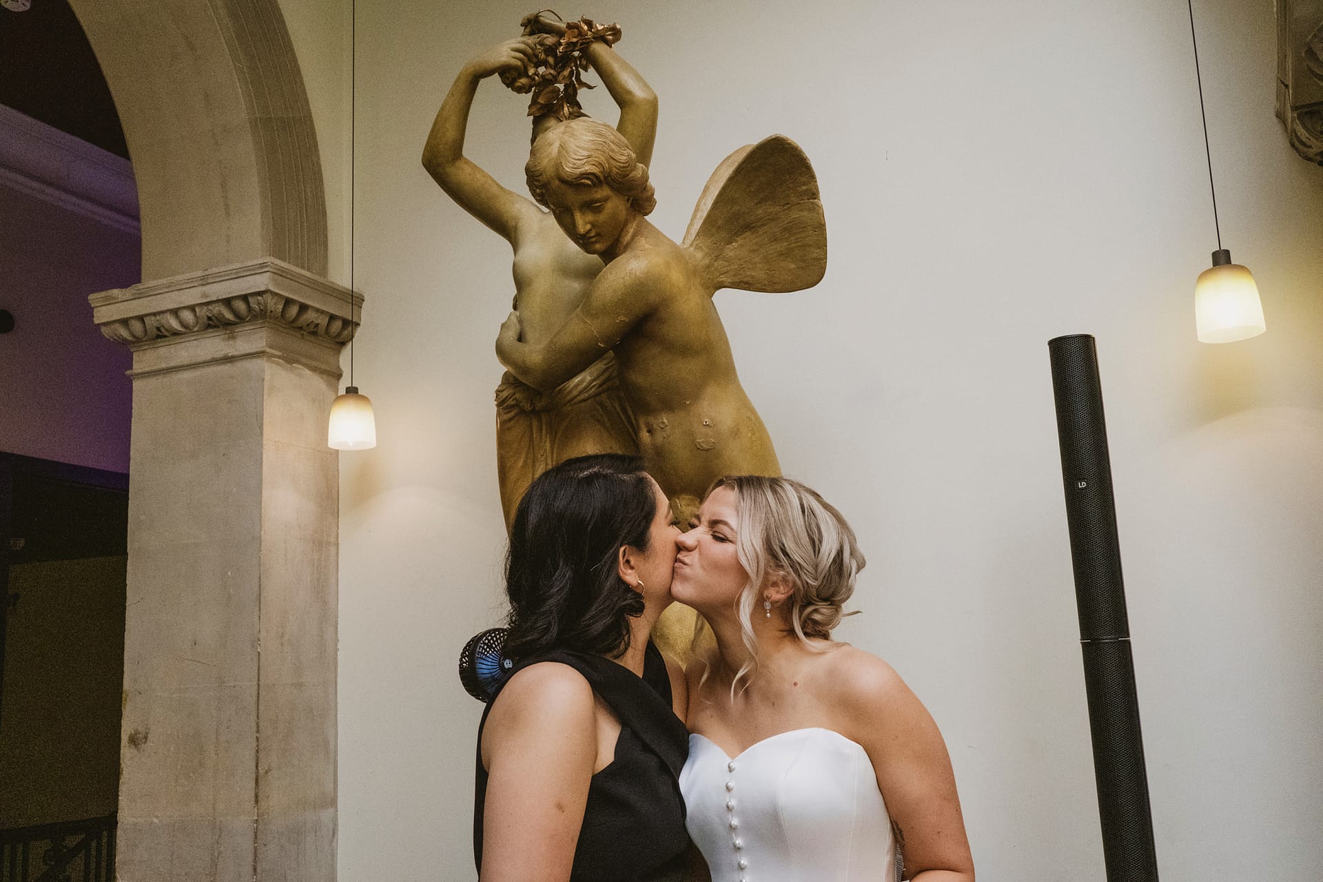 Two women kissing under a golden statue.