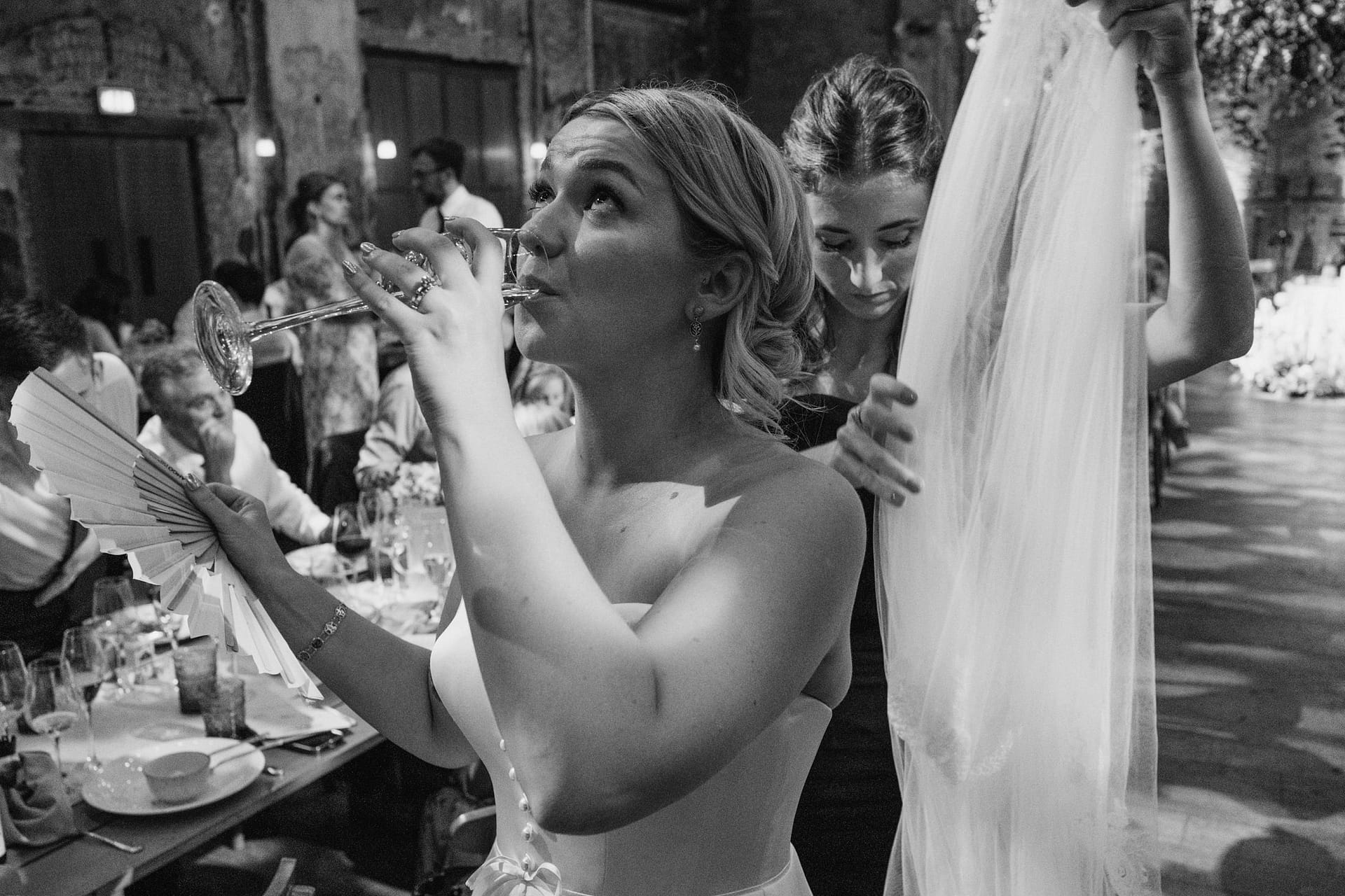 Bride drinking with fan and veil at wedding.