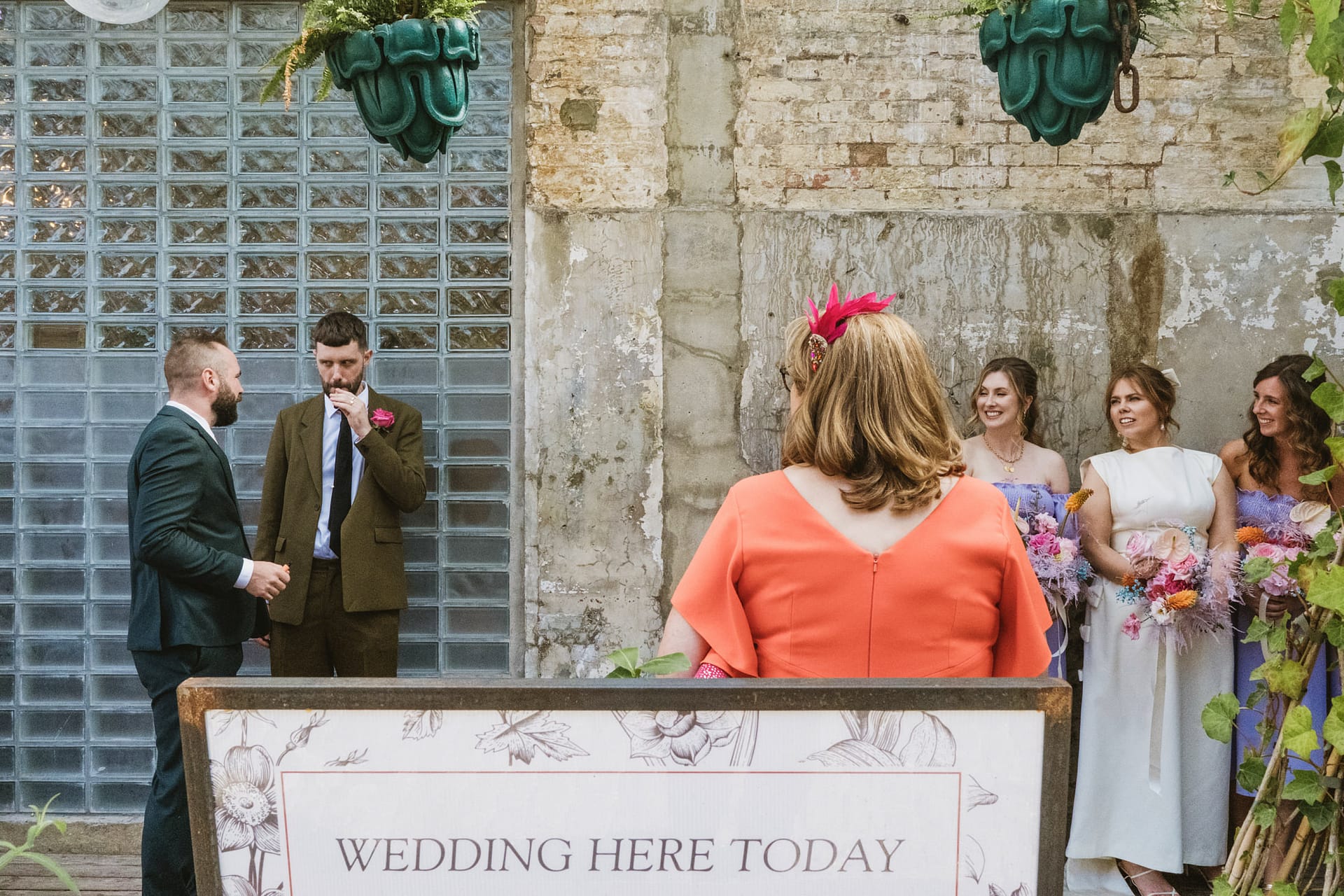 Outdoor wedding ceremony with guests, sign, and brick wall.
