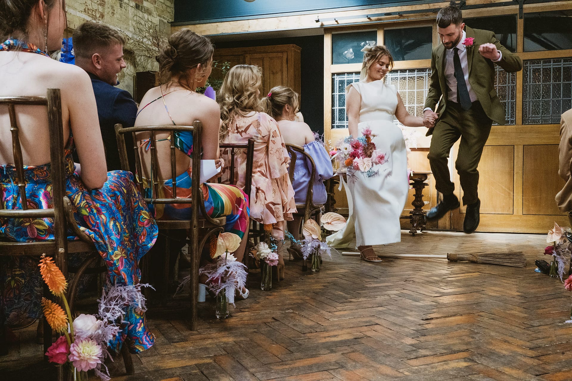 Wedding couple jumps over broom, guests watch.