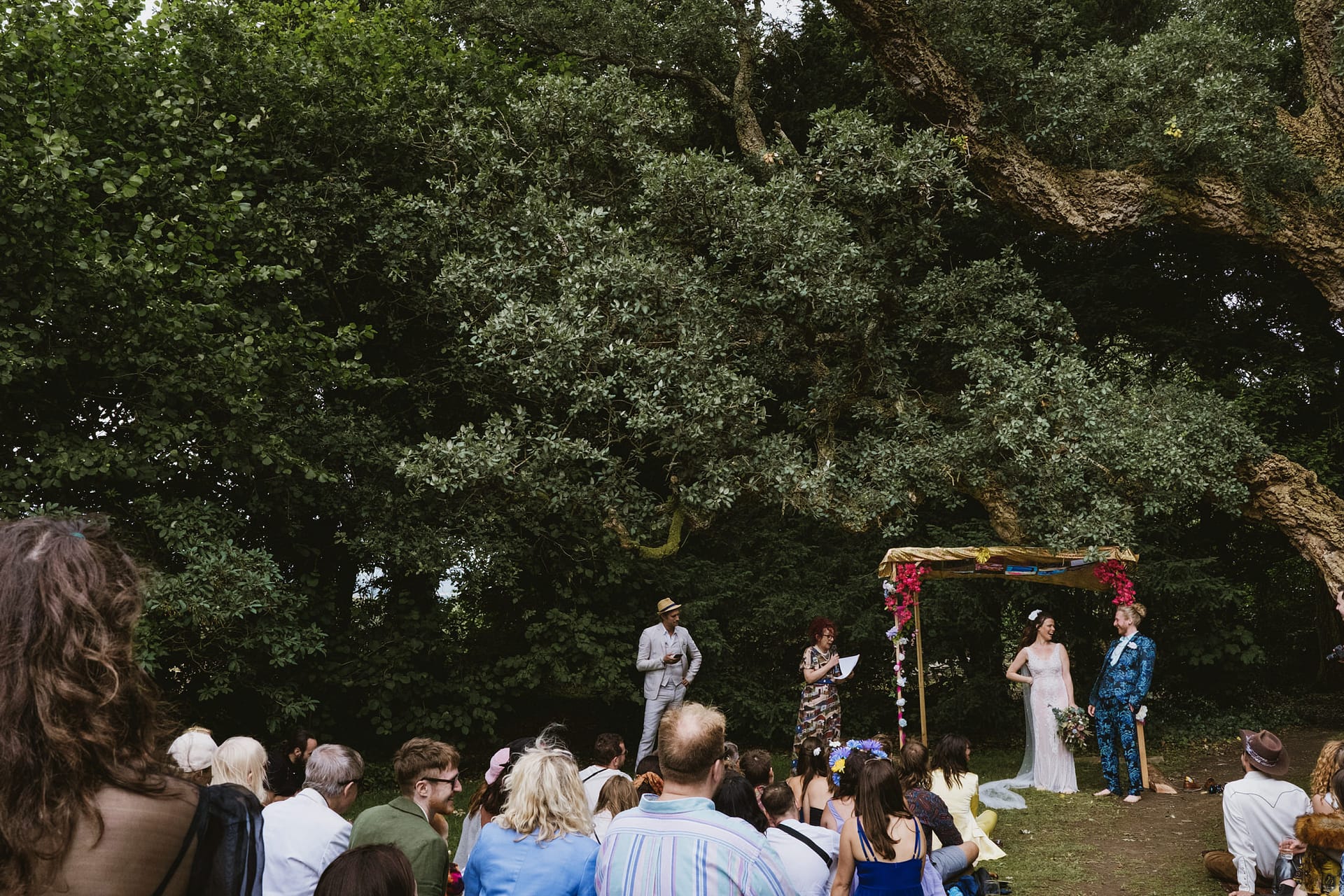 Outdoor wedding ceremony under tree with guests seated.