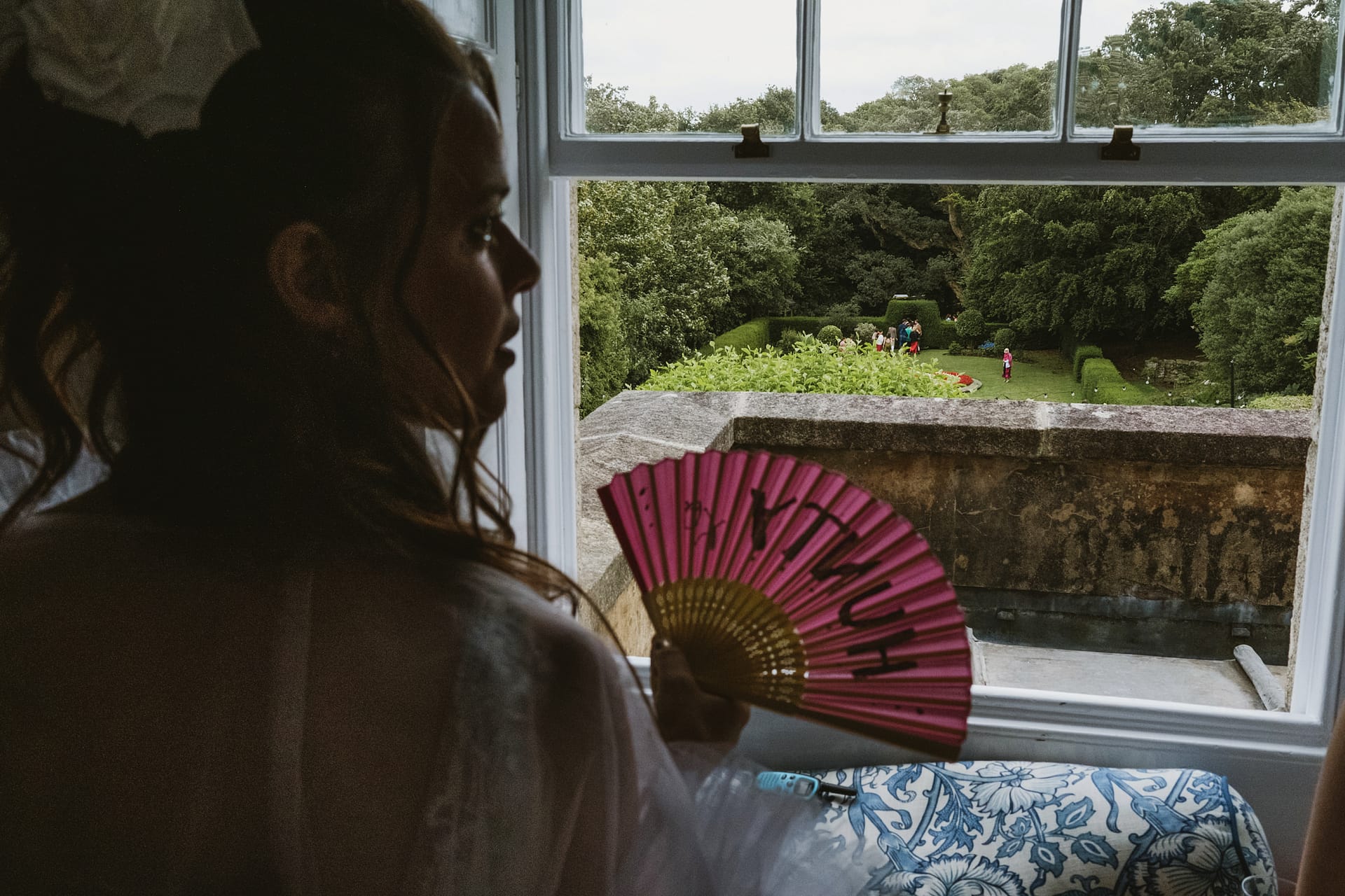 Bride holding fan, looking out window at the garden of Colehayes Park.