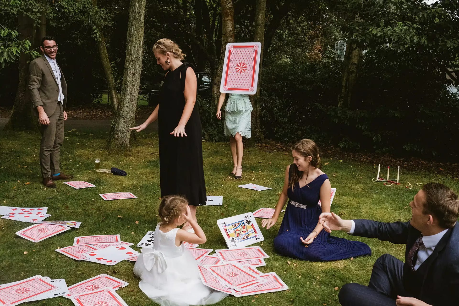 Layered, spontaneous documentary wedding photograph featuring 6 people on different planes of focus throwing oversized cards. One card (photographed flying through the air) covers the shoulders and head of a woman in the rear centre of the frame