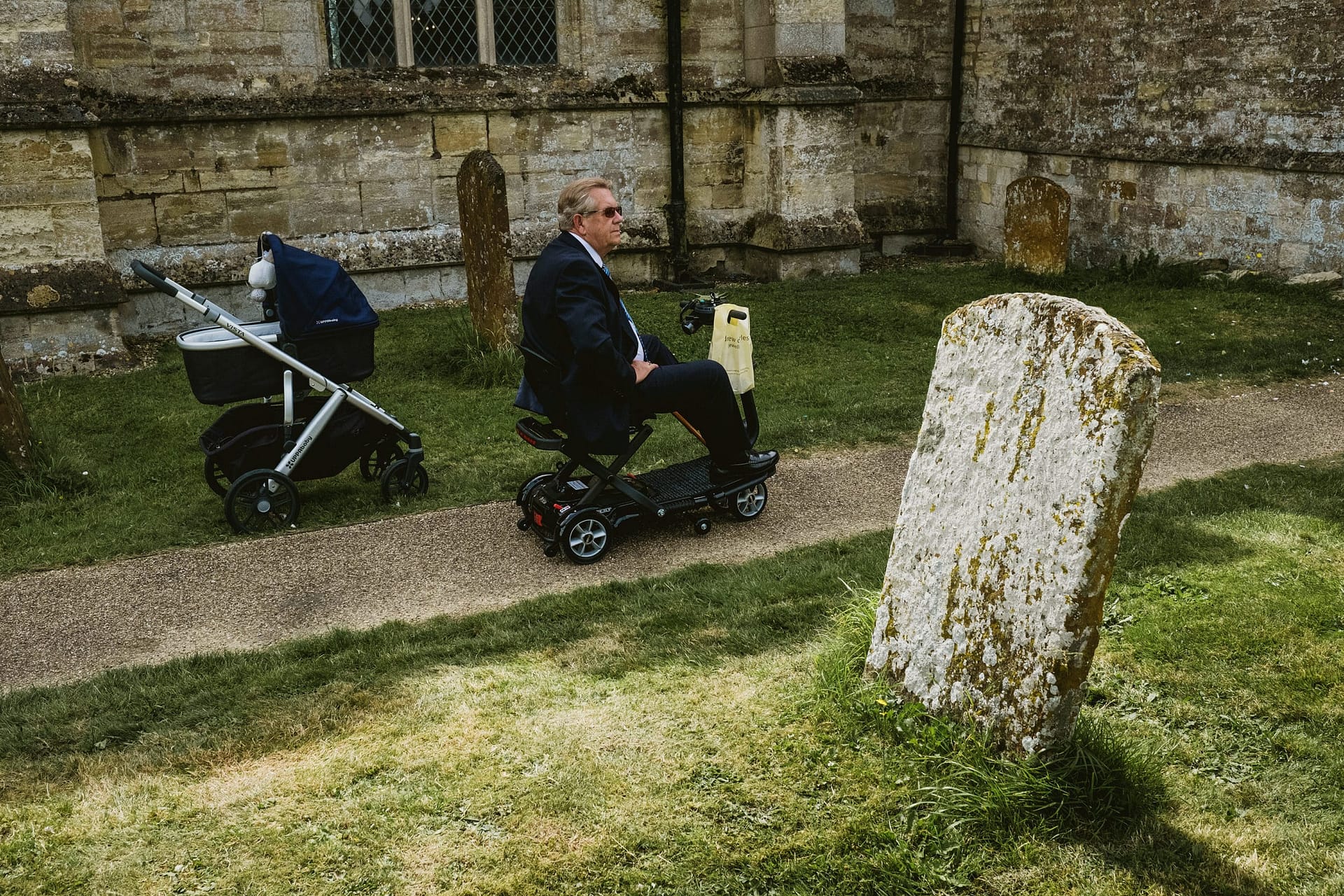 Documentary wedding photographer creates unconventional wedding story in the graveyard of a church. In the background on the left is a baby's pram, in the centre a man on a mobility scooter drives down the path and in the foreground (right) is a gravestone, depicting "the journey of life from cradle to the grave"