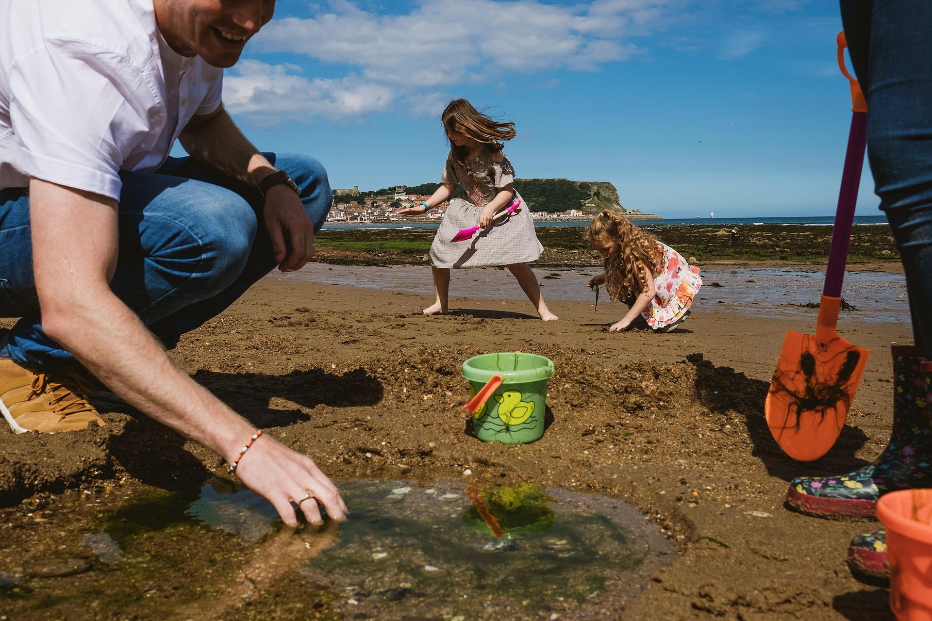 Family Day Out photography on a beach by York Place. A Man is crouched down looking in a rock pool on the left of the image. Next to him is a bucket and opposite someone wearing wellington boots is standing holding a spade with a crab painted on it. In the centre of the frame in the background two girls are playing.