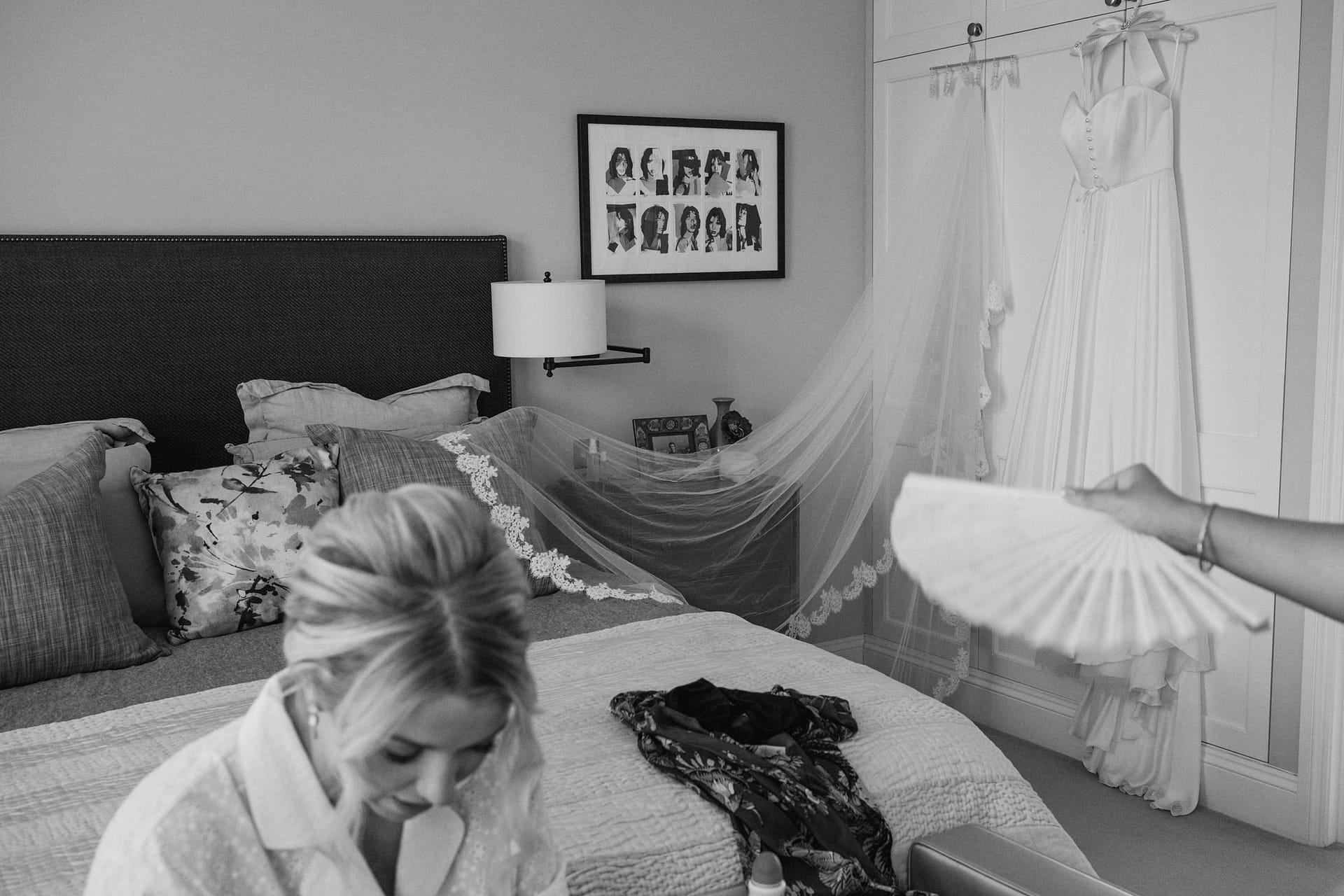Bride preparing in bedroom, wedding dress hanging nearby.