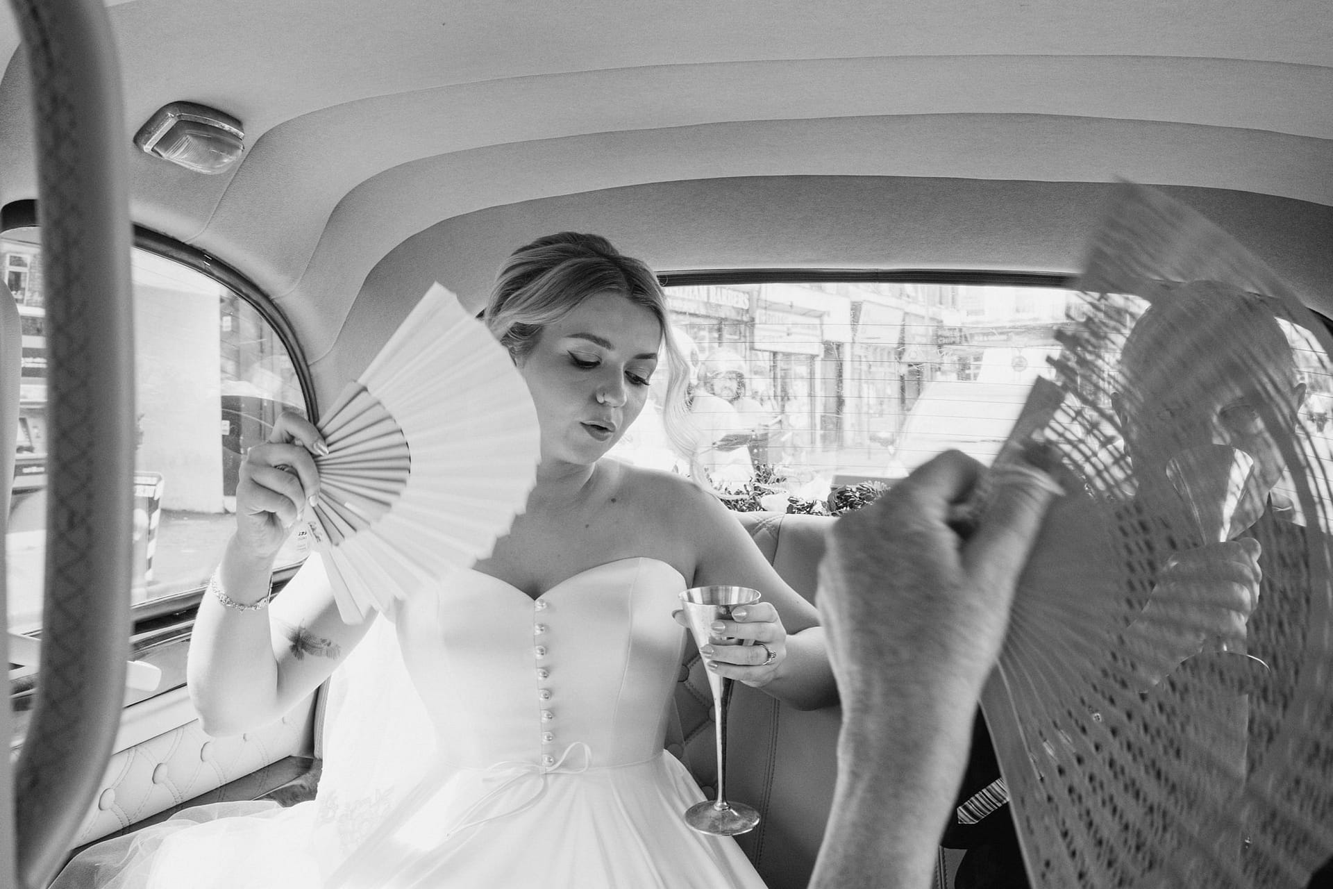 Bride using fan and holding drink in car.