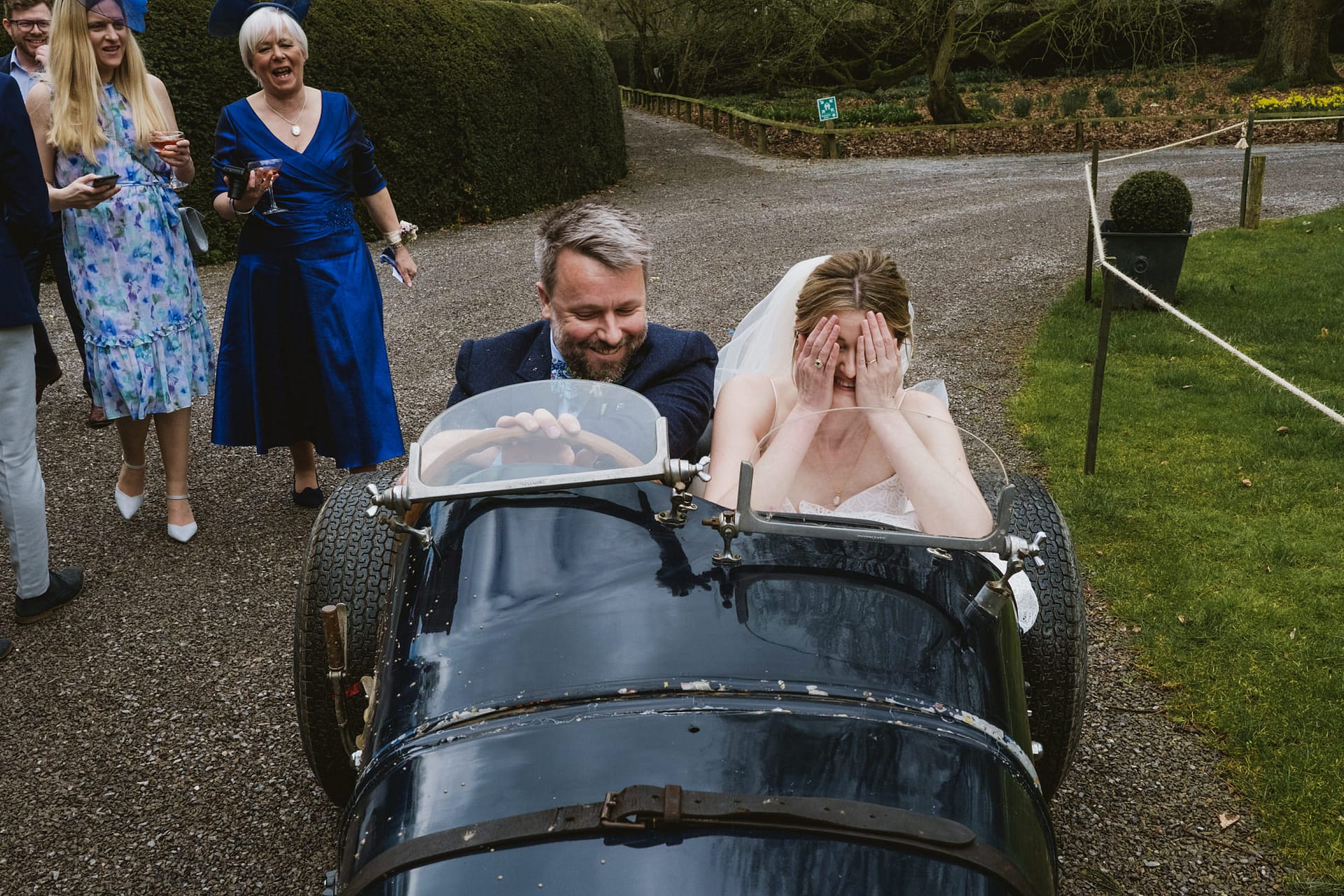 Bride covers face in vintage car, guests laughing.