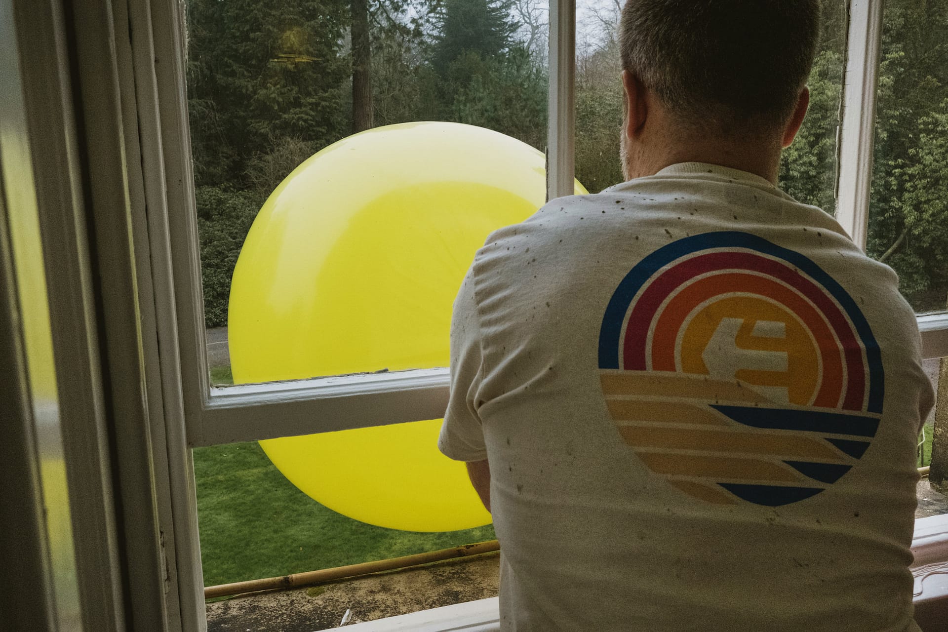 Man inflating large yellow balloon outside window.