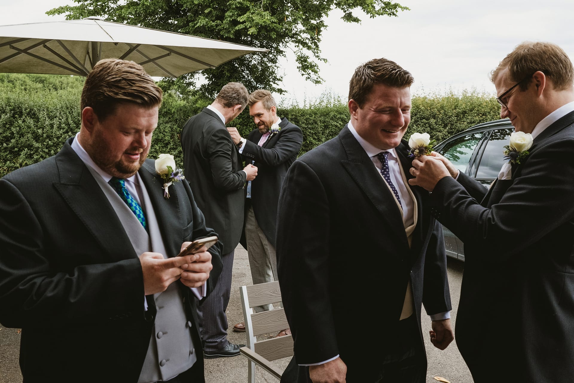 The groom has his button hole put on by his groomsmen whilst in the background another groomsmen has his button hole pinned at the pub The Chequers inn in Buckinghamshire.