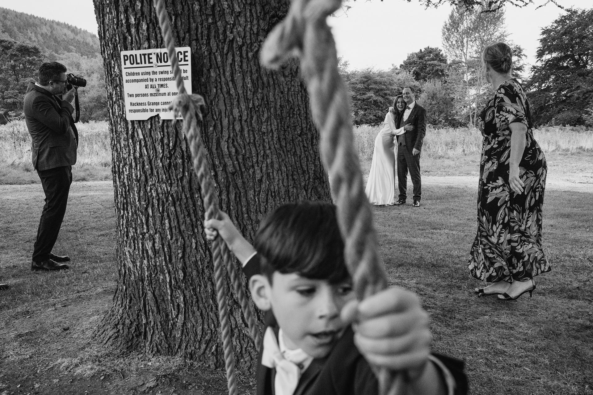 Candid wedding moment - photograph showing guests relaxing during wedding reception, framed around a tree. A child is on a swing in the foreground with a guest to the left. To the right is another guest with a camera, looking at the bride and groom in the background