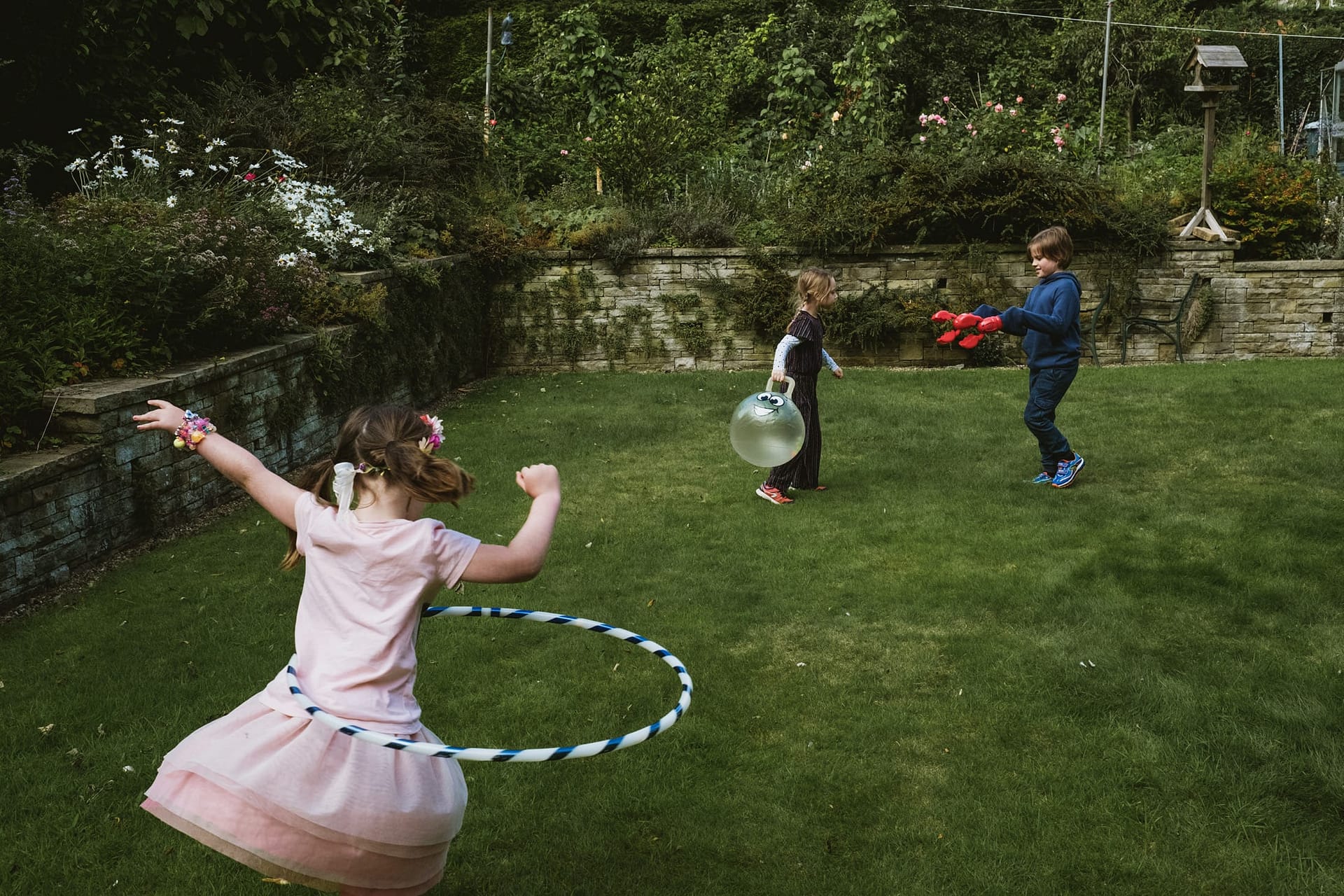 family portrait photography captured in a family garden - young girl in pink dress spins a hula hoop in the foreground, whilst in the background another girl holds a spacehopper as her brother approaches her with lobster claw toys replacing his hands