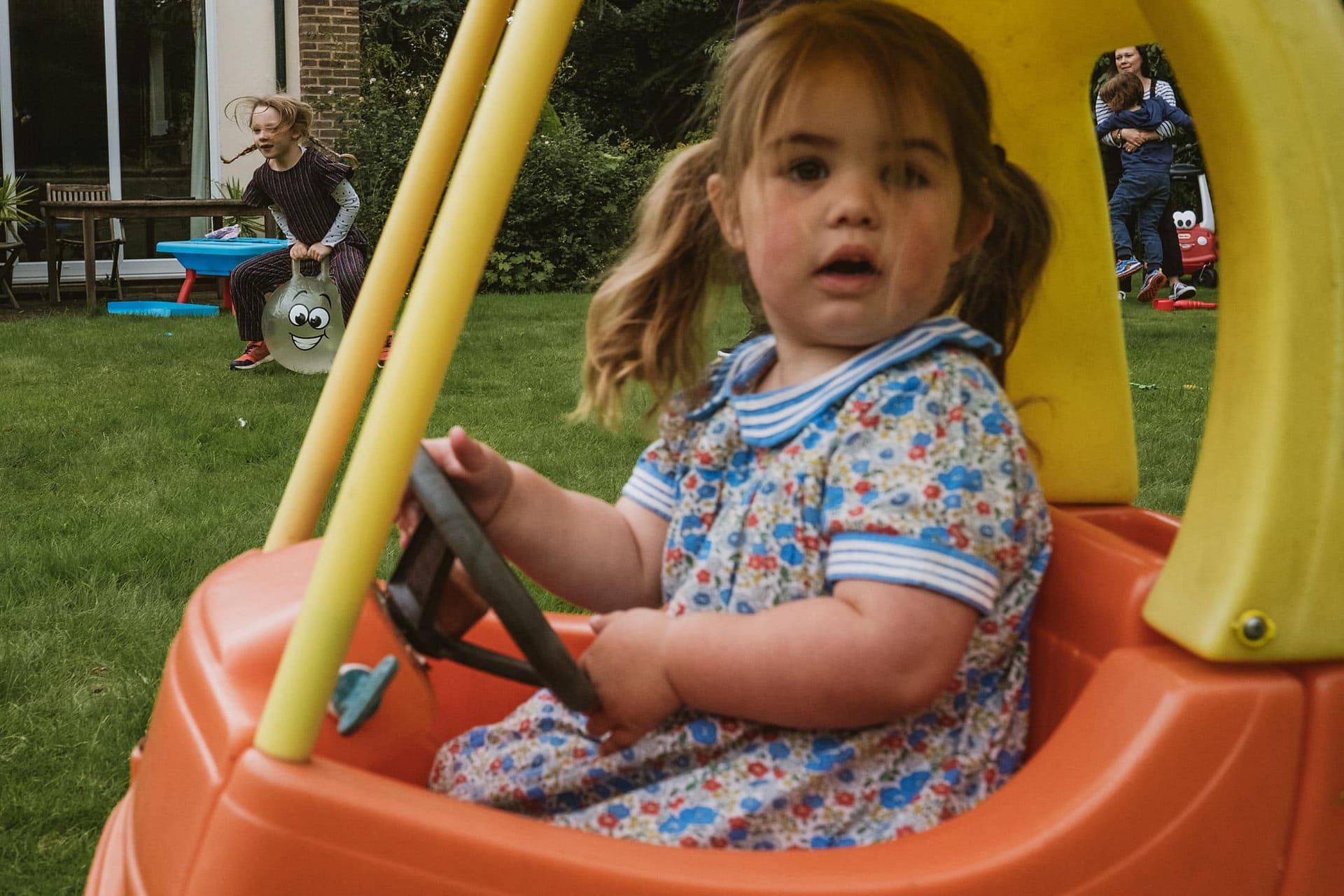 little girl in toy car and other children in the background. Documentary family photography