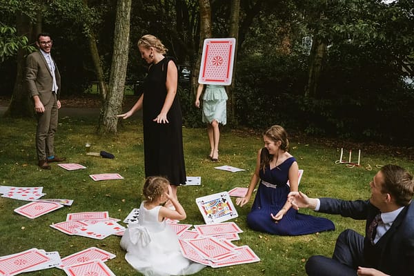 Layered, spontaneous documentary wedding photograph featuring 6 people on different planes of focus throwing oversized cards. One card (photographed flying through the air) covers the shoulders and head of a woman in the rear centre of the frame
