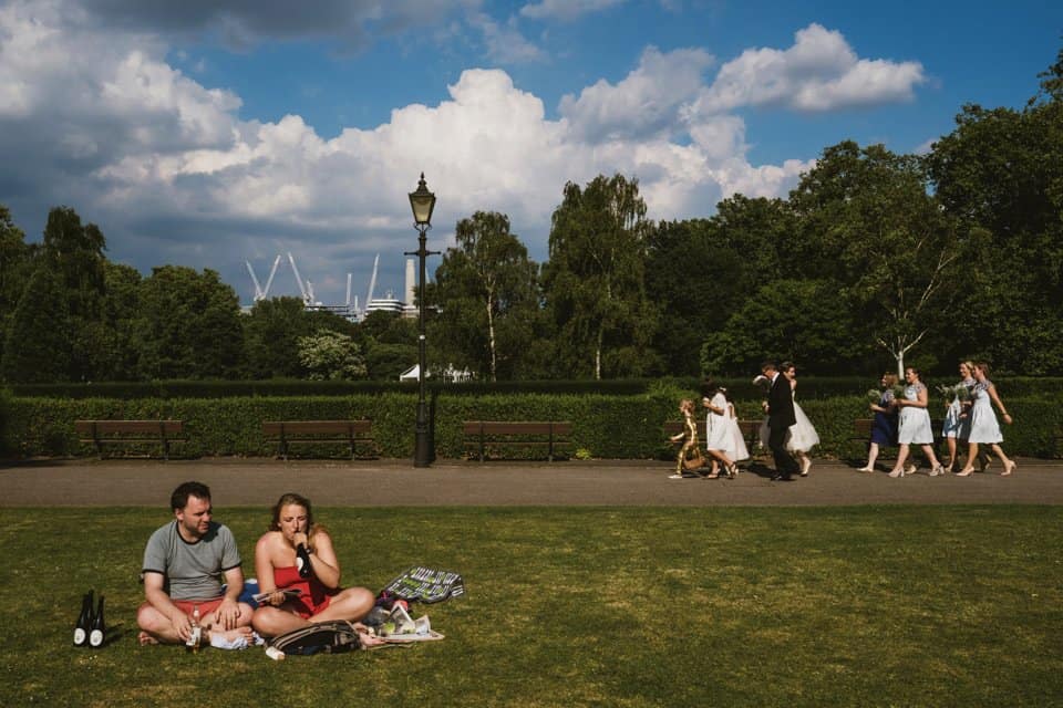 London wedding photographers Dominique & Liam Shaw capture an alternative take on the walk to the wedding venue, including a picnic (with wine bottle in hand) in the foreground whilst the wedding party walk by in the background in front of the London skyline