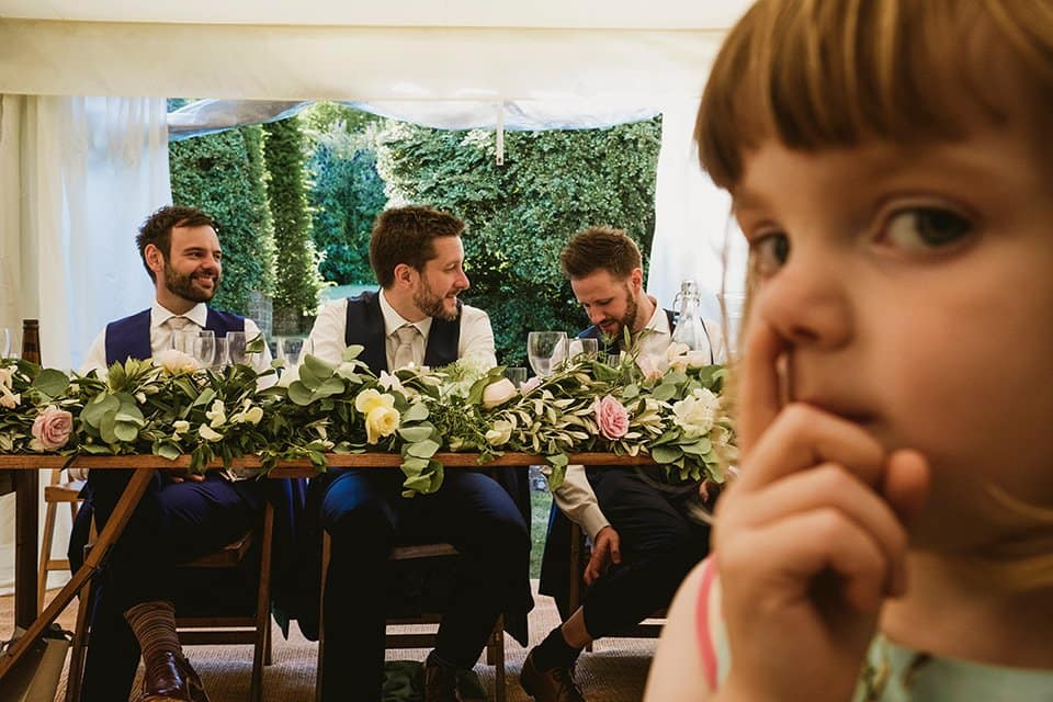 Comedic wedding photograph - as three groomsmen sit at the top table in the background, to the right and in the foreground a young girl appears to pick her nose close to the camera